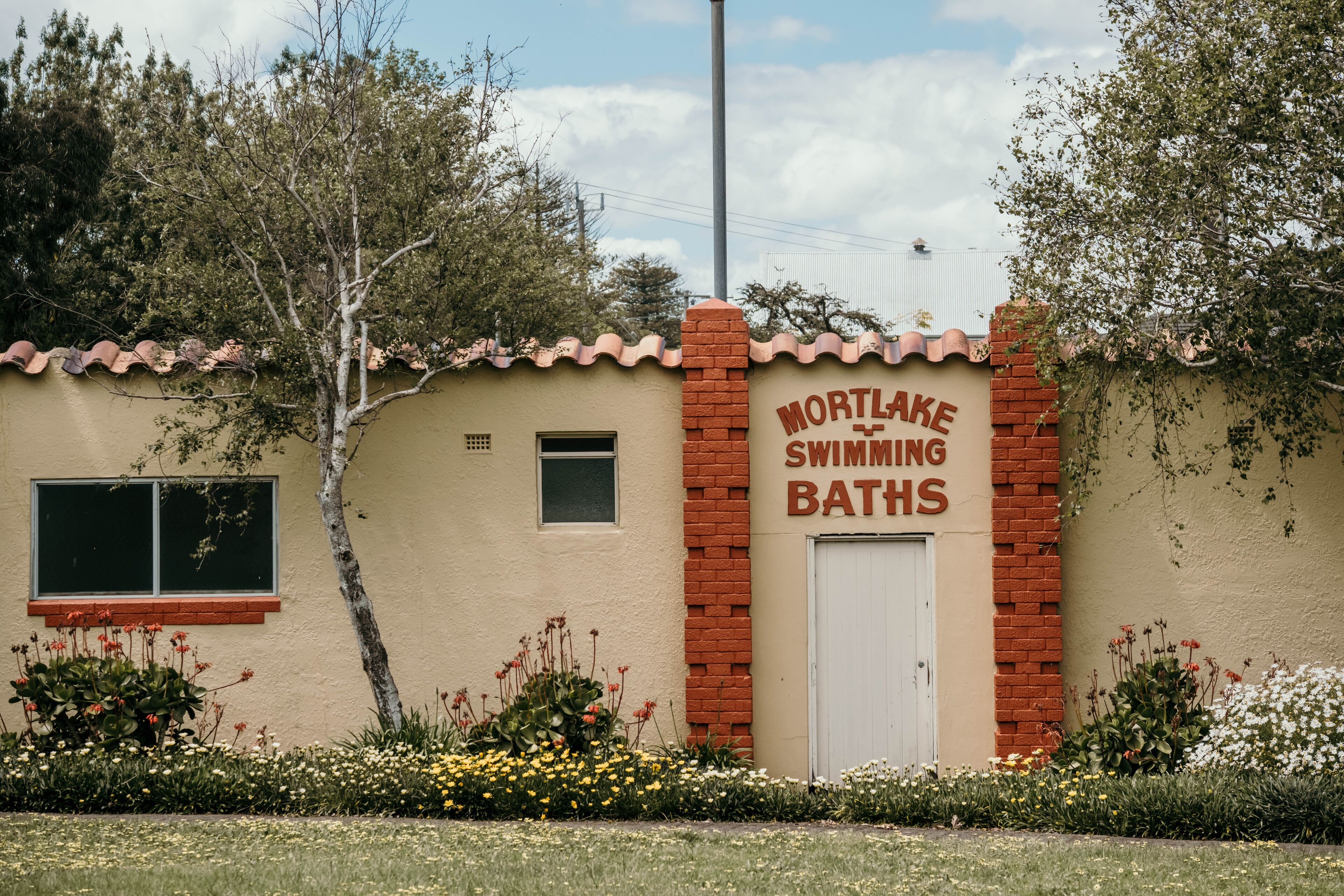 A building with a sign reading "Mortlake Swimming Baths" and a red brick entrance.