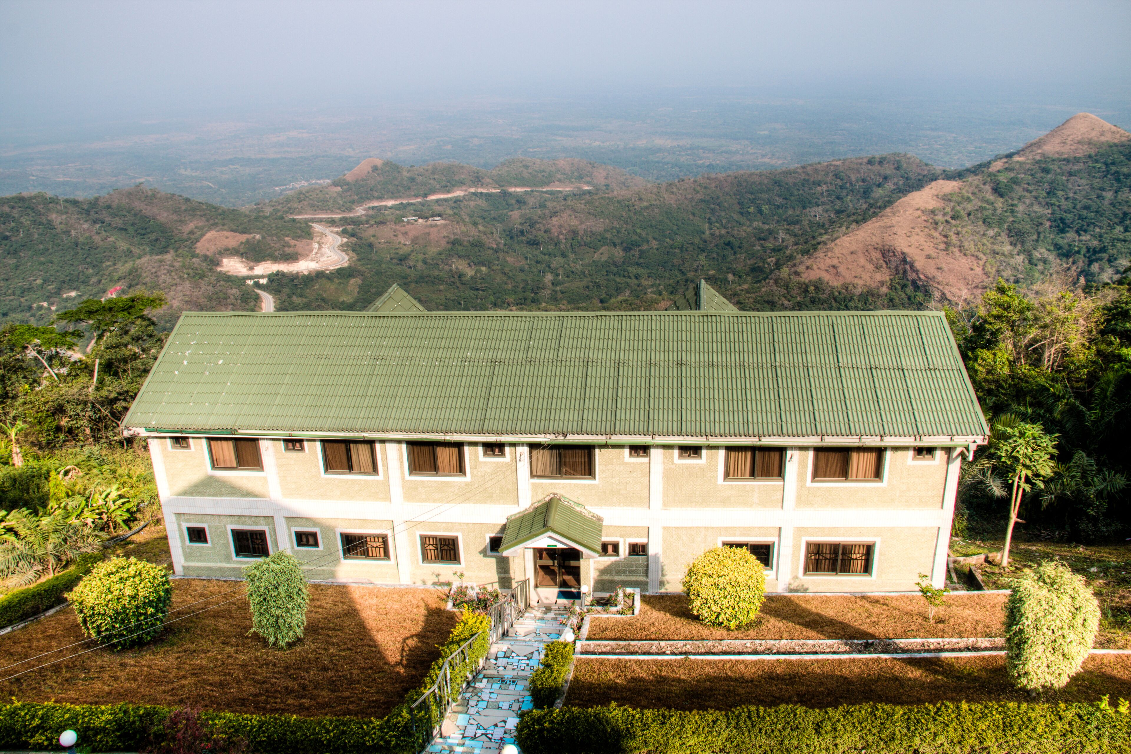 A large two-story building with a green roof, surrounded by lush greenery and hills in the background.