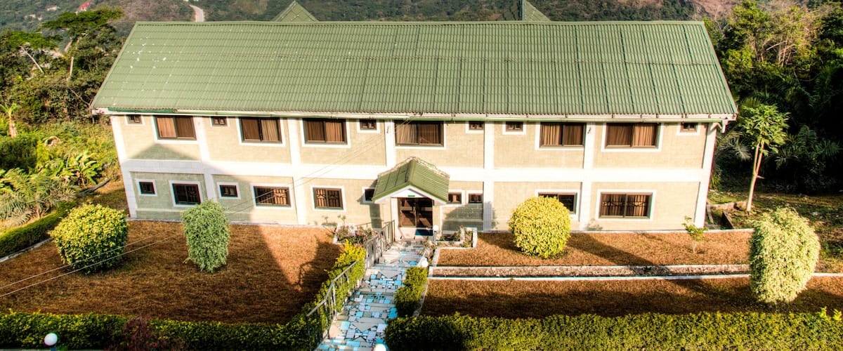 A large two-story building with a green roof, surrounded by lush greenery and hills in the background.