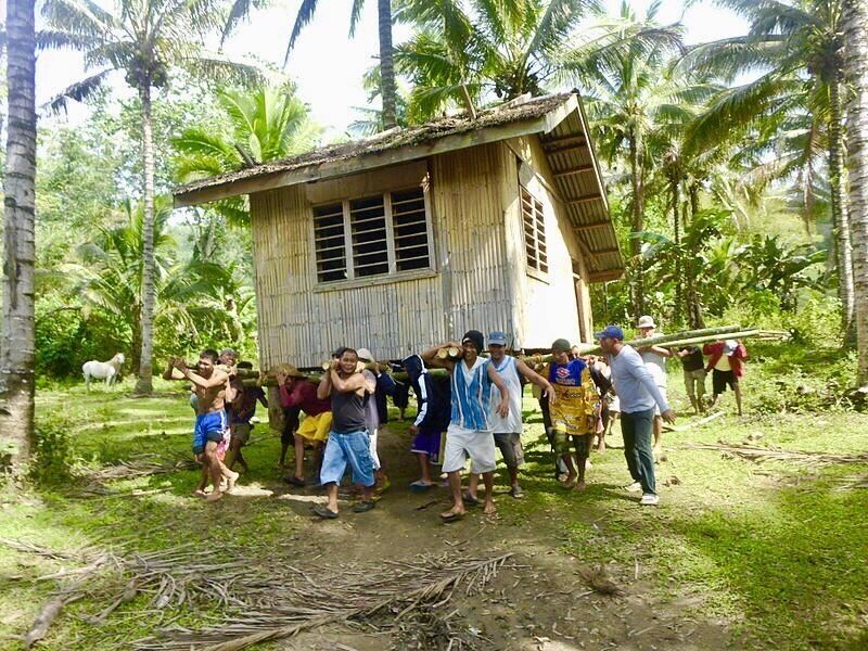 A group of people standing in front of a wooden house in a tropical setting with palm trees.