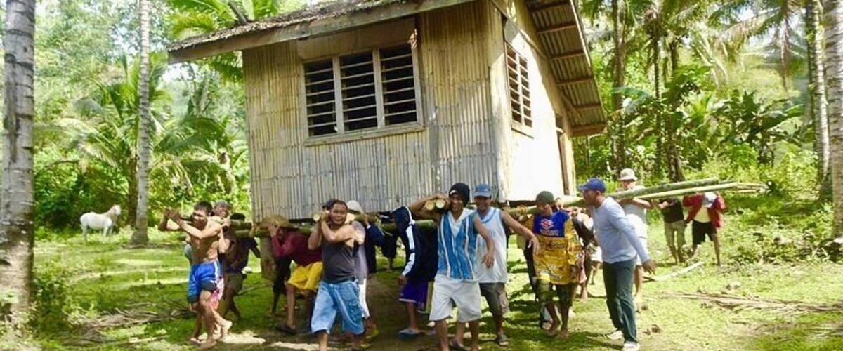 A group of people standing in front of a wooden house in a tropical setting with palm trees.