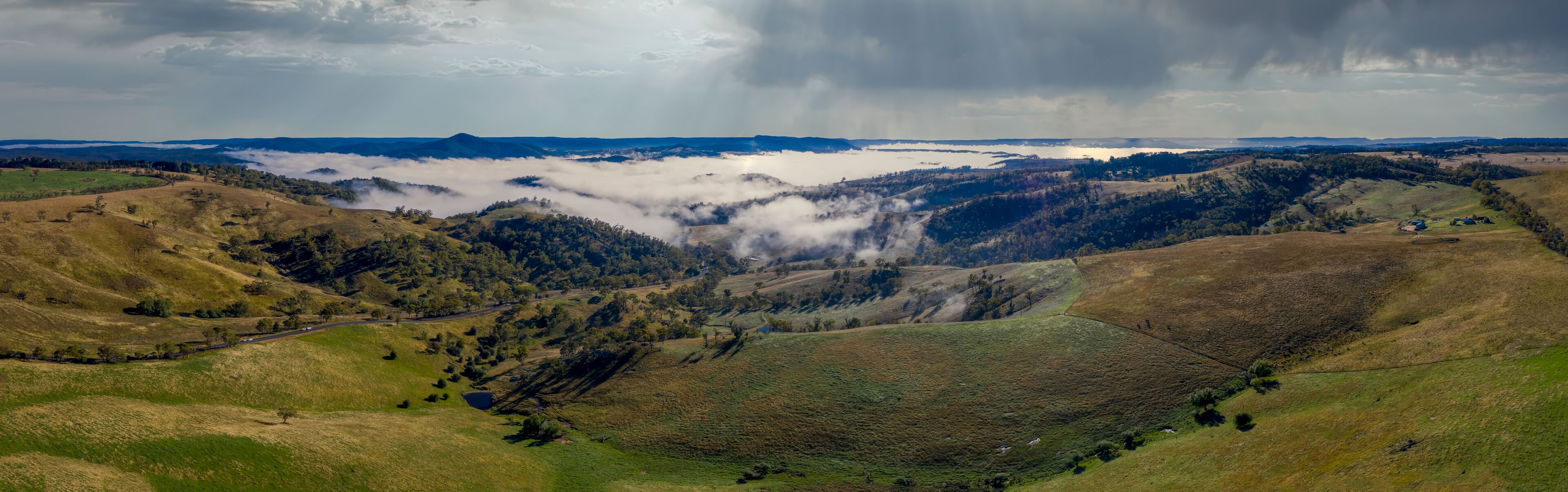 A valley with rolling hills, a lake, and a mountain range under a cloudy sky.