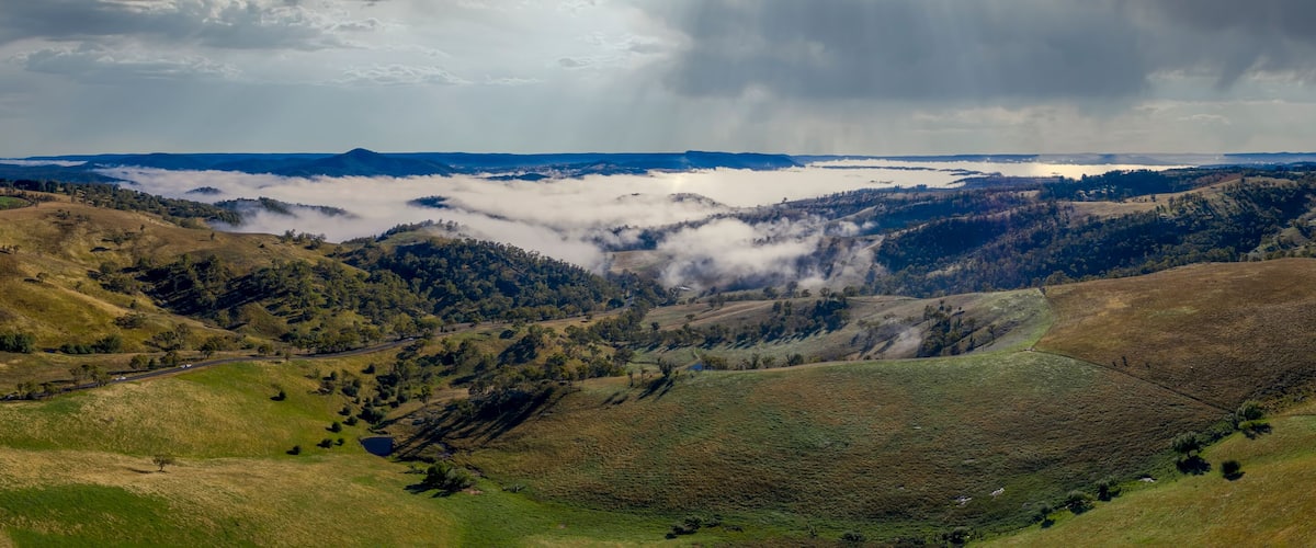 A valley with rolling hills, a lake, and a mountain range under a cloudy sky.