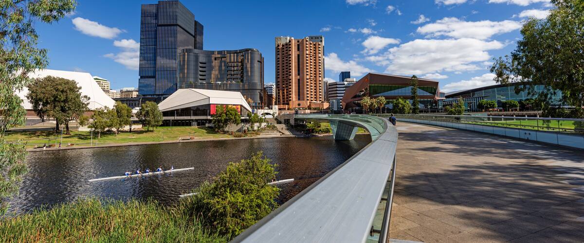A river with a bridge, modern buildings, and a park with trees.
