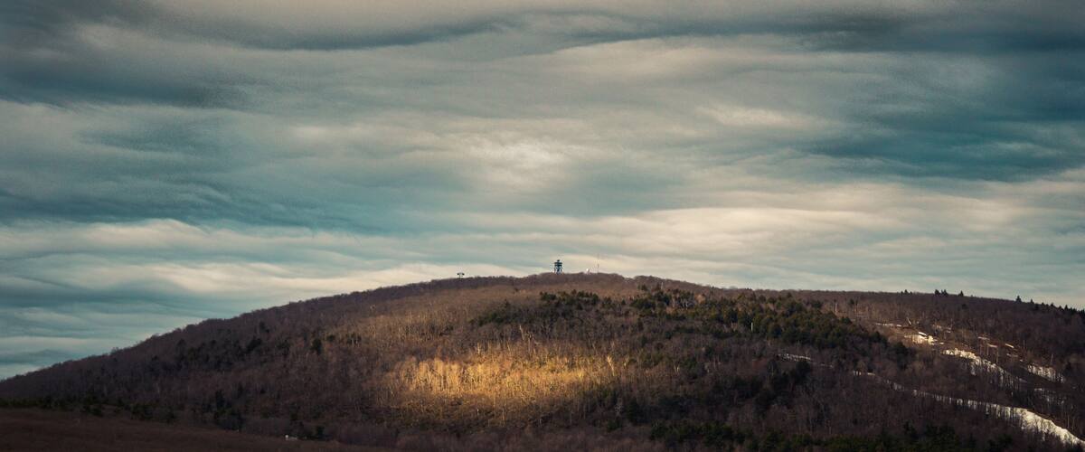 A hilly landscape with a tower on top, dense forest, and a cloudy sky.