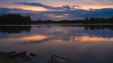 A calm lake at sunset with a reflection of the sky, surrounded by trees and rocks.