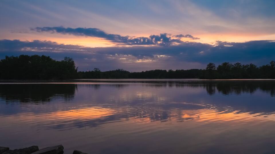 A calm lake at sunset with a reflection of the sky, surrounded by trees and rocks.