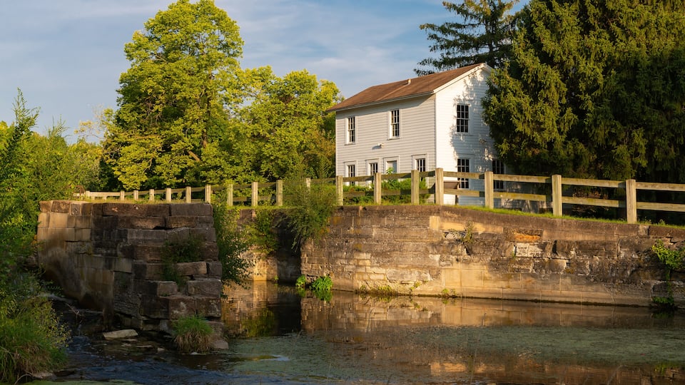 A stone bridge over a calm river, surrounded by lush greenery and a white house with a wooden fence.