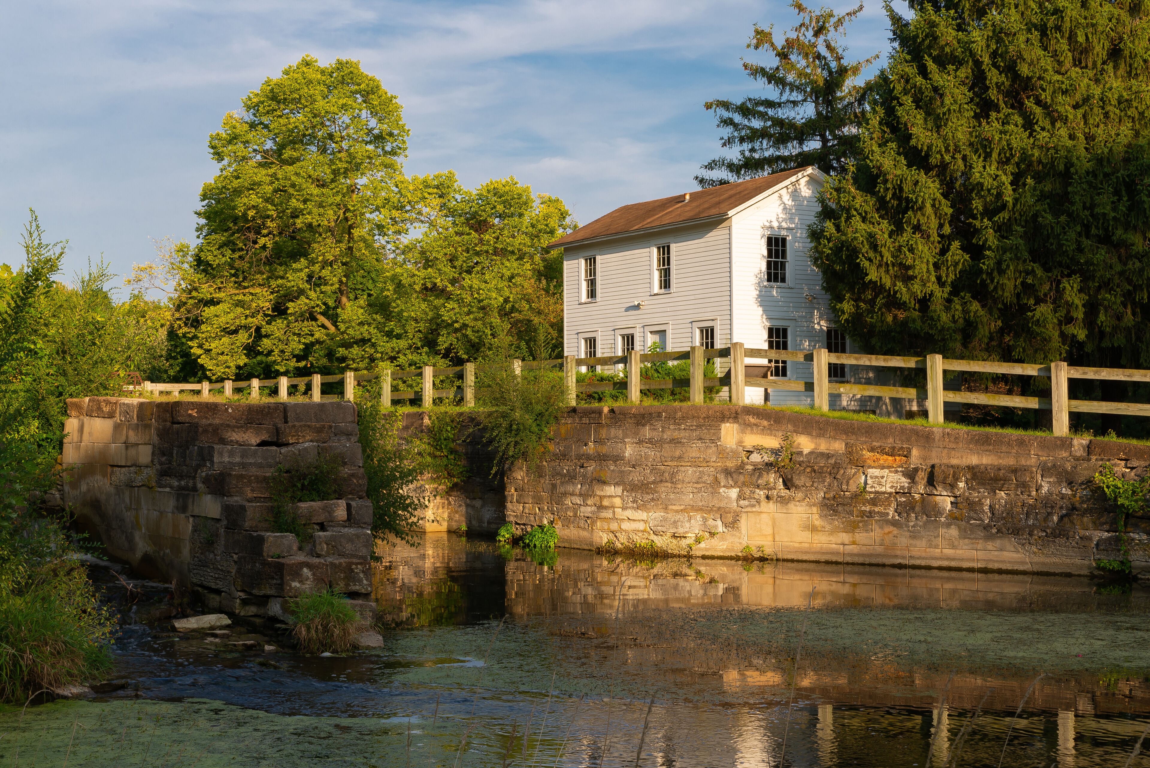 A stone bridge over a calm river, surrounded by lush greenery and a white house with a wooden fence.
