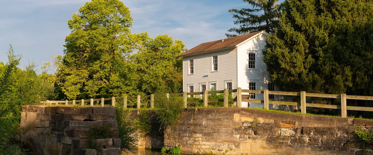 A stone bridge over a calm river, surrounded by lush greenery and a white house with a wooden fence.