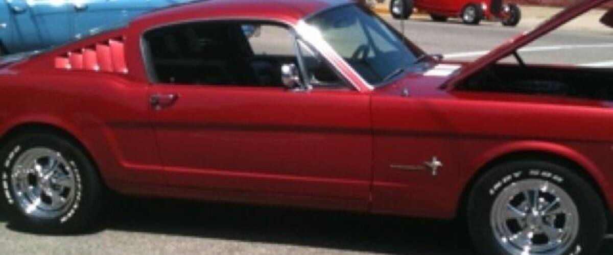 A red vintage car with its hood open, parked on a street with other vintage cars and a crowd of people.