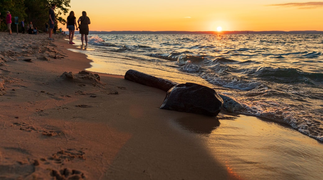 A beach with people walking along the shore, waves crashing onto the sand, and a sunset in the background.