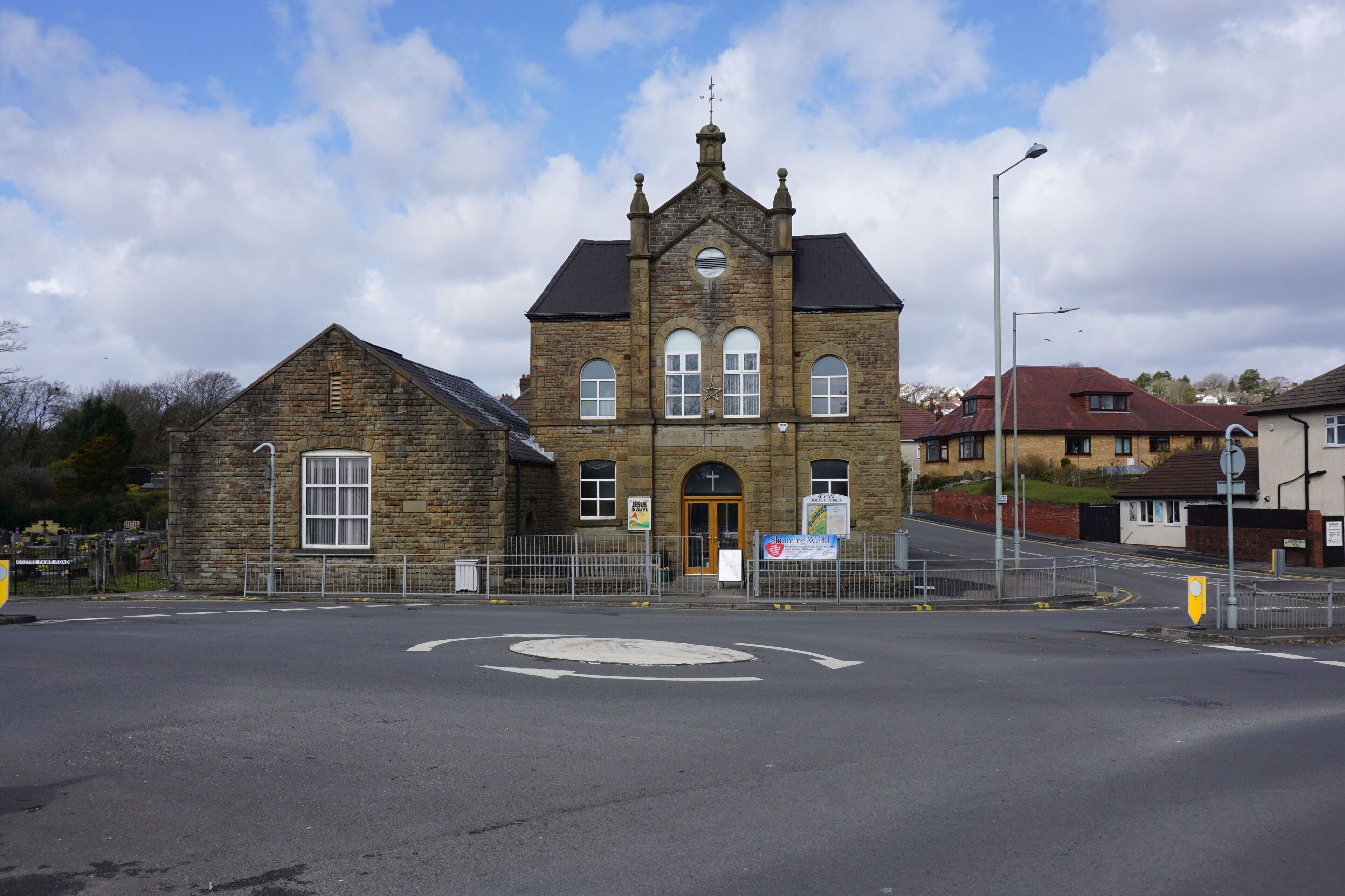 A historic stone building with a clock tower, a roundabout, and a street with houses in the background.
