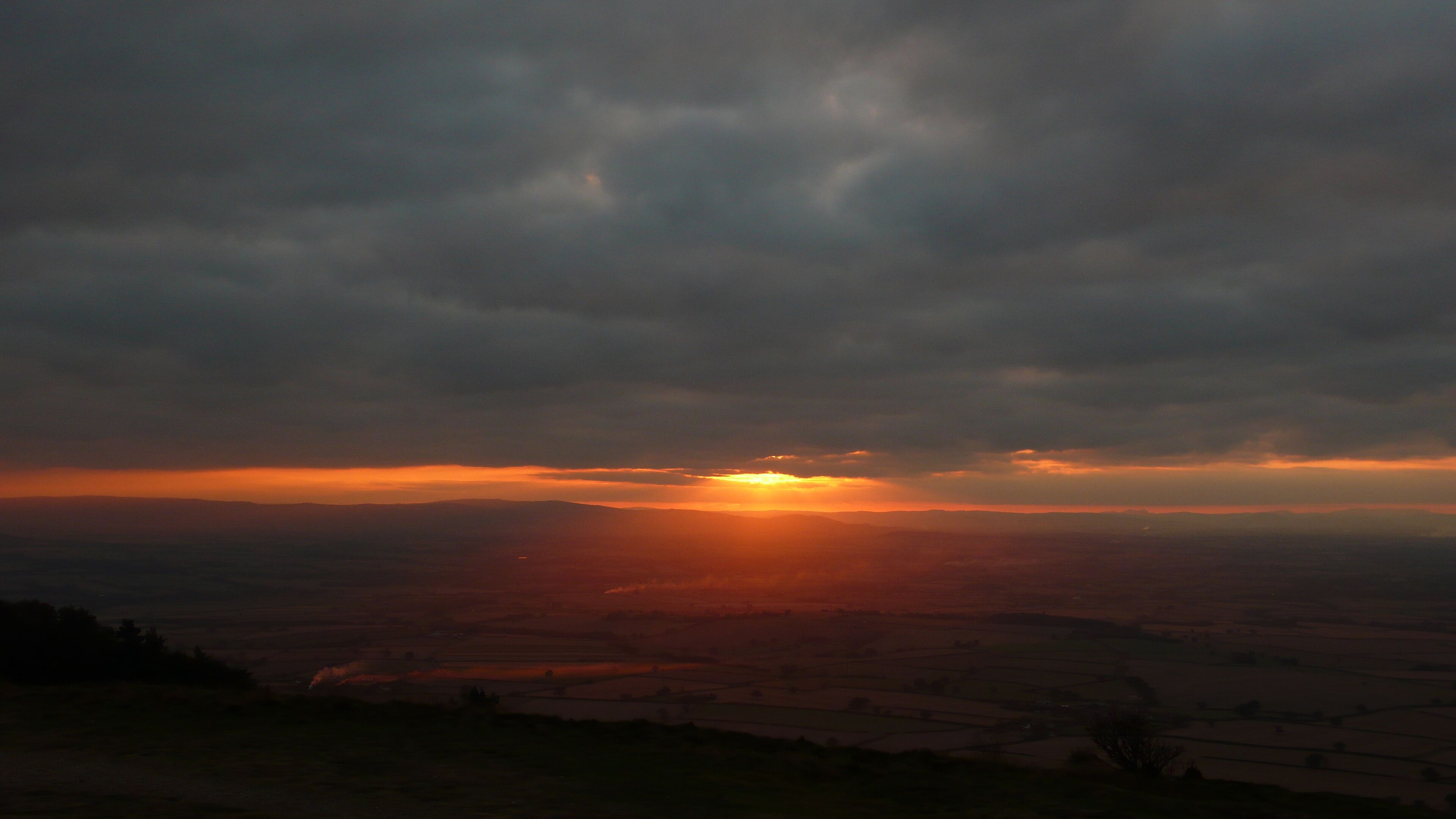 A sunset over a rural landscape with fields and a cloudy sky.
