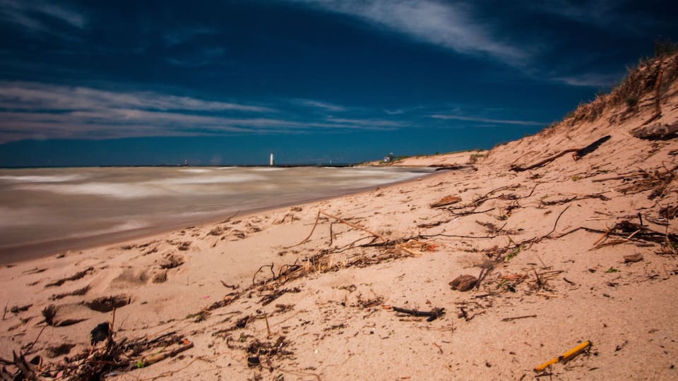 A sandy beach with scattered driftwood and a lighthouse in the distance.