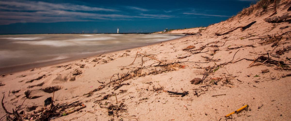 A sandy beach with scattered driftwood and a lighthouse in the distance.
