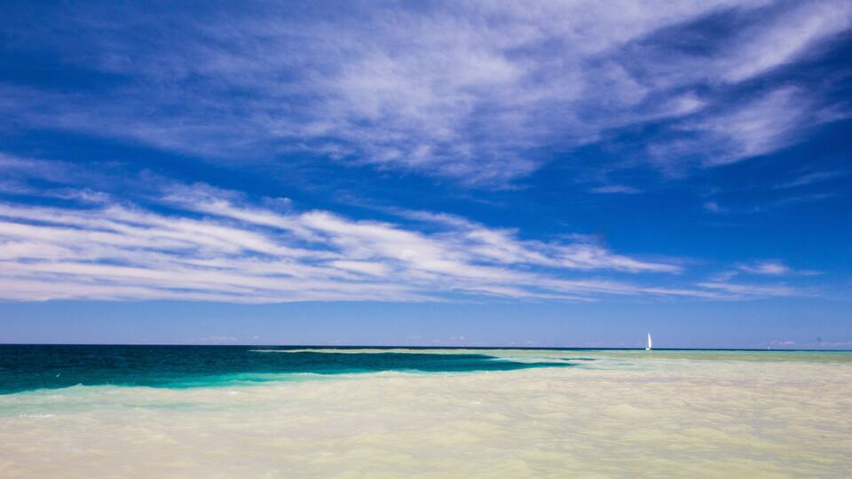 A beach with clear turquoise water, a sandy shore, and a sailboat in the distance.