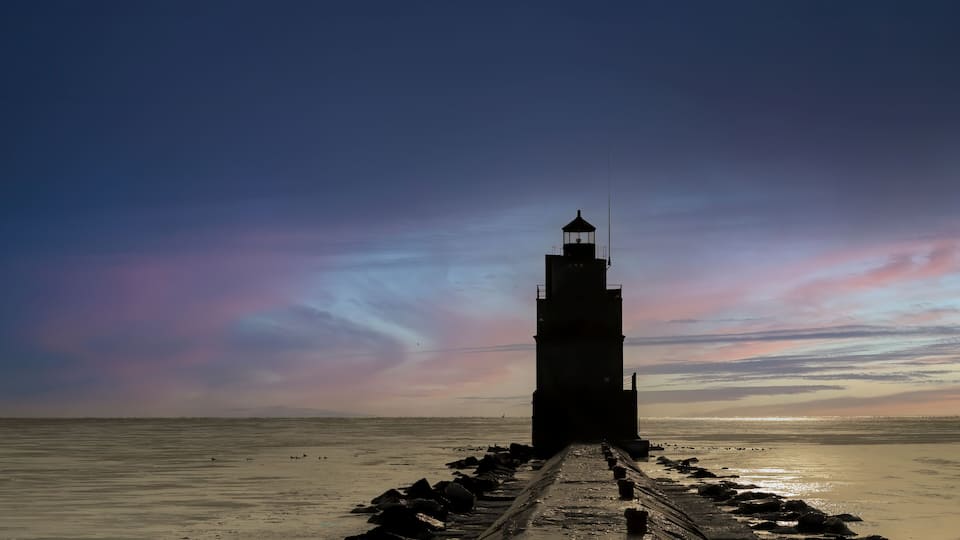 A lighthouse on a pier extending into the sea with a sunset sky.