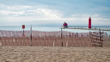 A family, just visible through fence slats, enjoys a walk on the beach, at Grand Haven, MI, lighthouse and pier in background.