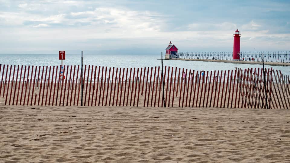 A beach with a wooden fence, a red lighthouse, and a pier with a small house.