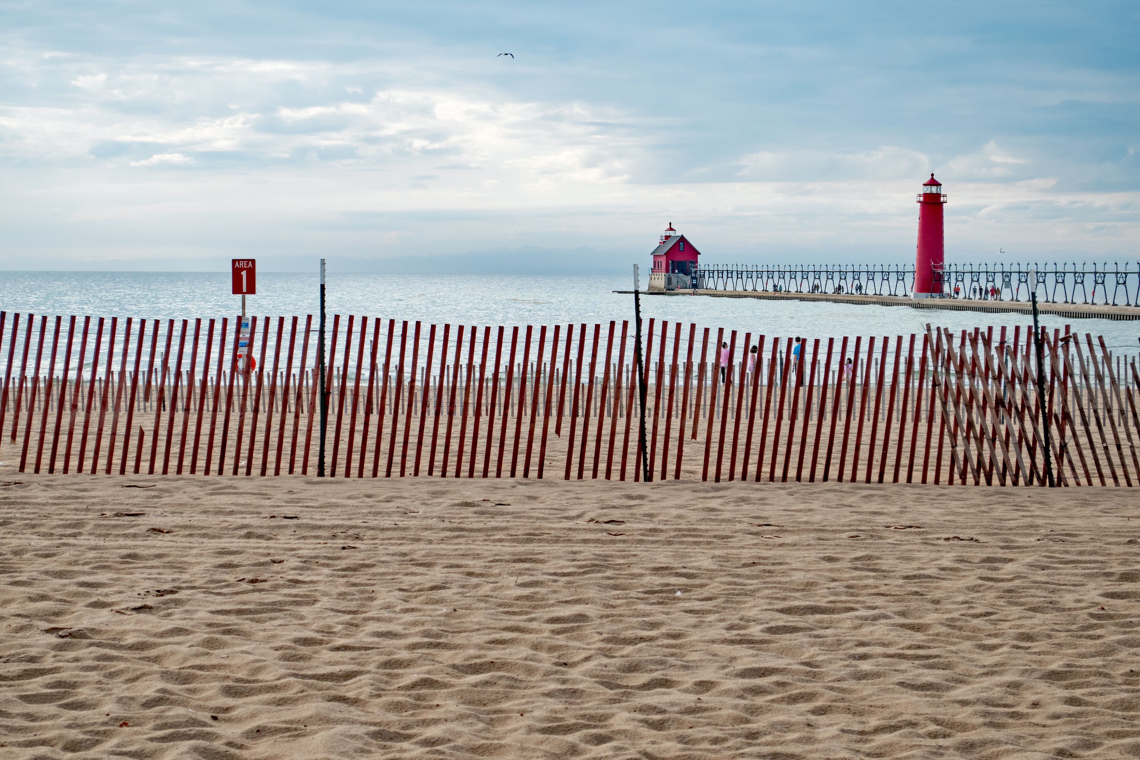A beach with a wooden fence, a red lighthouse, and a pier with a small house.