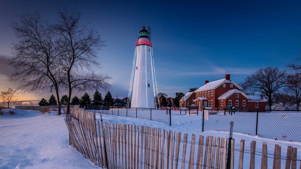 A snow-covered landscape with a lighthouse, a wooden fence, and a brick house.