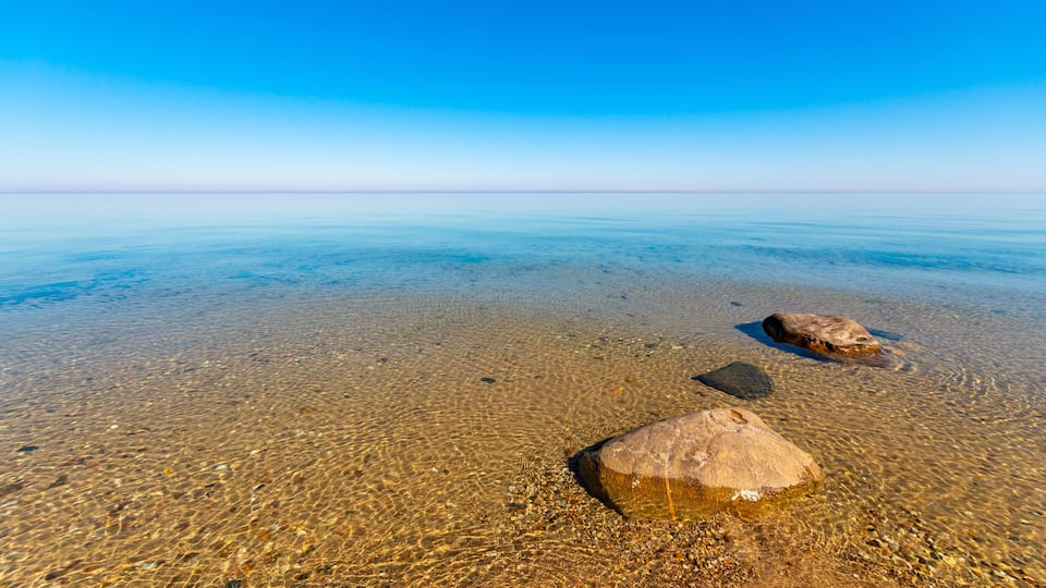 A clear blue sky, calm sea, and two large rocks on a pebbly beach.