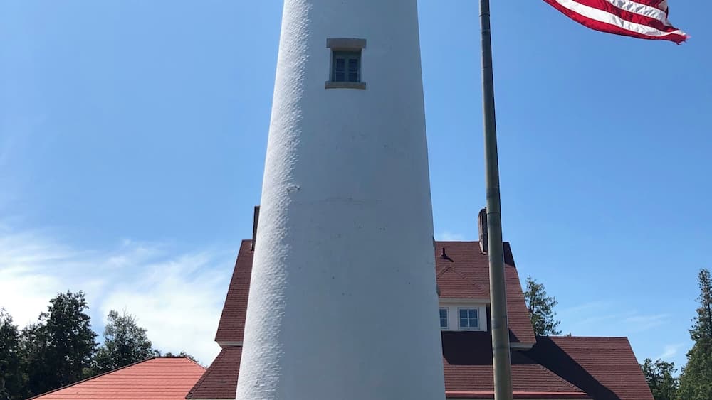 A white lighthouse with a red roof, an American flag, and a brick house with a red roof in the background.