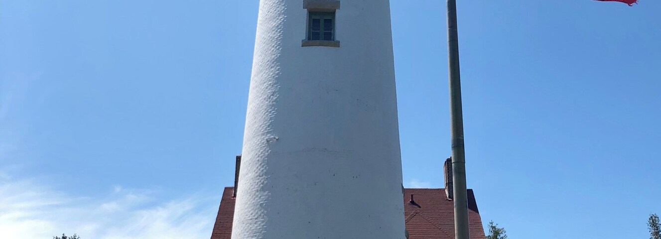 Un faro blanco con techo rojo, una bandera estadounidense y una casa de ladrillo con techo rojo al fondo.