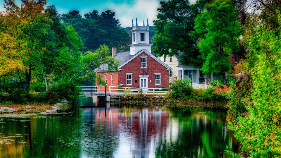 A red brick building with a white tower, surrounded by lush greenery and a calm river.