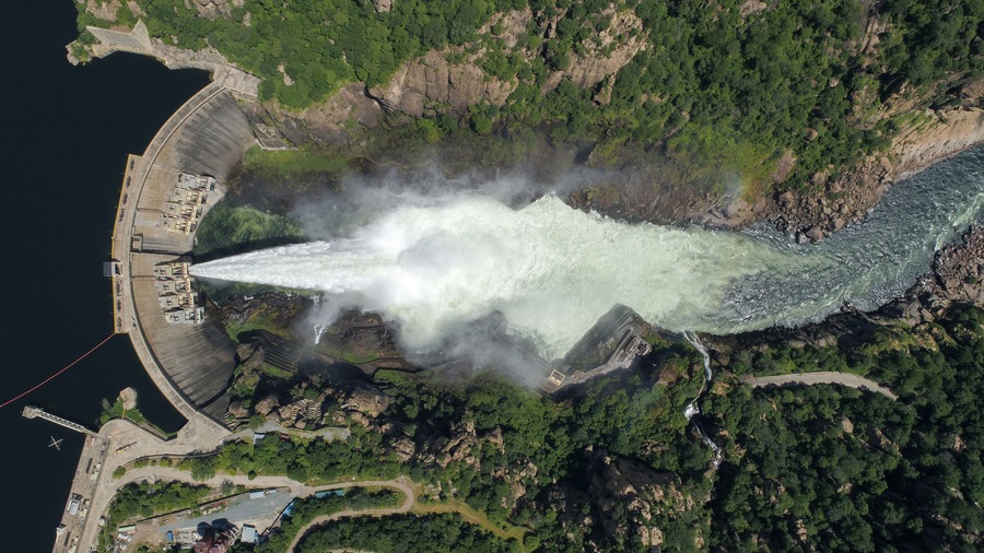 A dam with a large water spill, surrounded by greenery and a river.