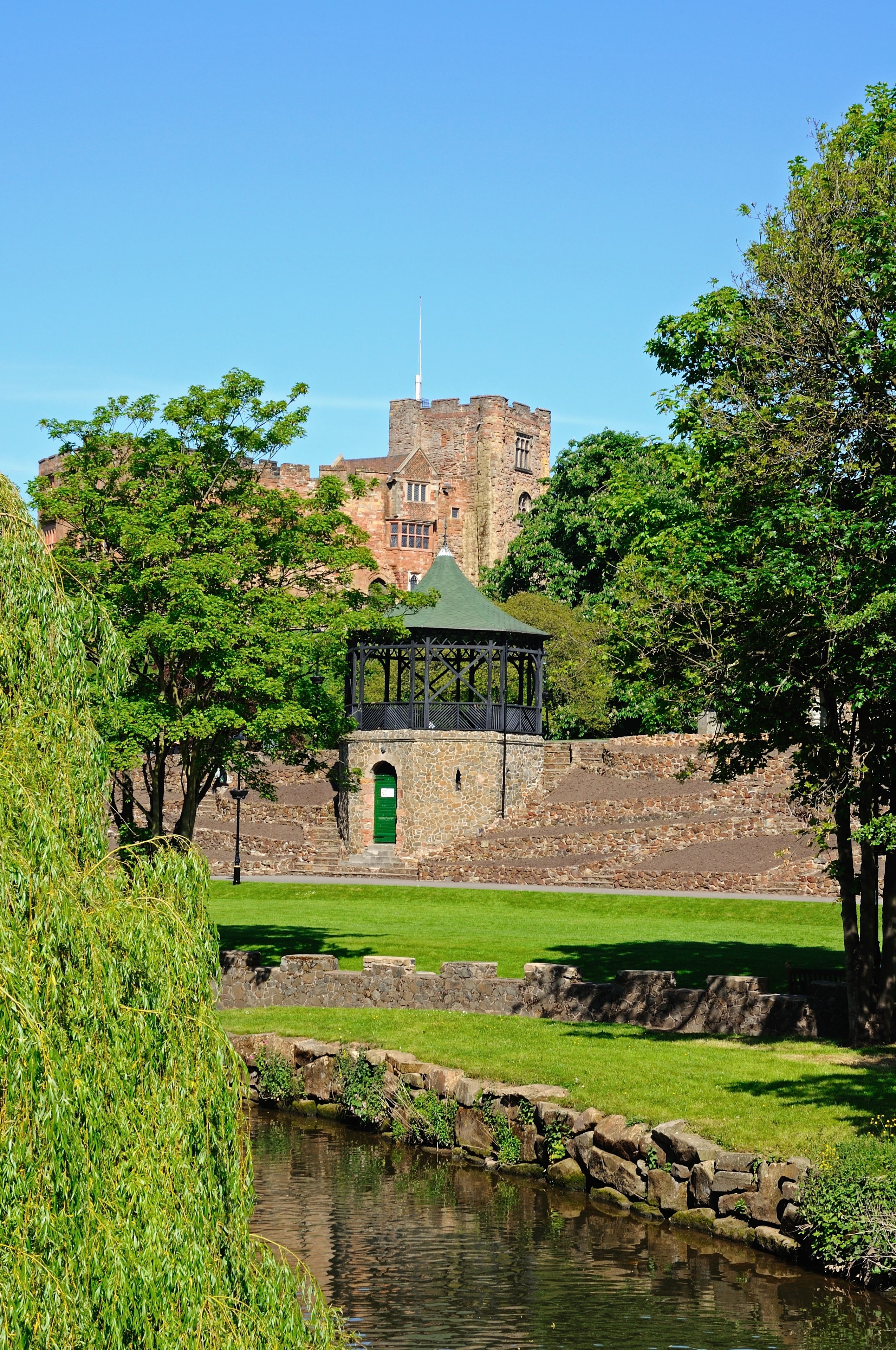A stone structure with a green roof, a small pond, and a large castle in the background.