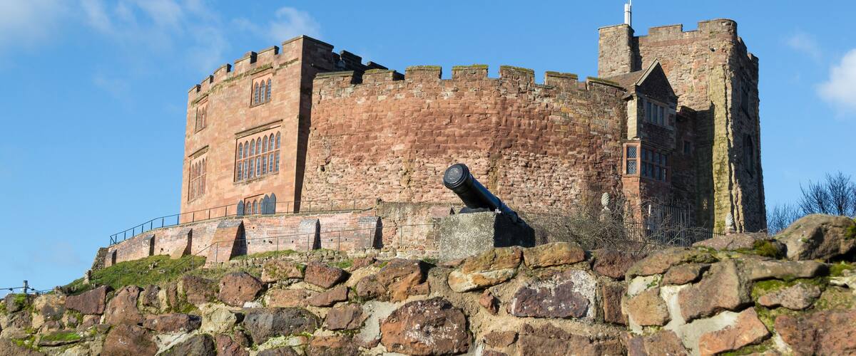 Un castillo de piedra histórico con una bandera en la parte superior, un cañón grande y un cielo azul despejado.