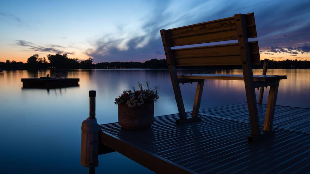 A wooden bench on a dock with a potted plant, overlooking a calm lake at dusk.