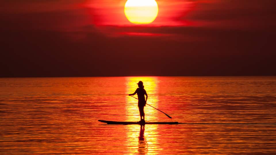 A silhouette of a person paddleboarding at sunset with a large sun reflecting on the water.