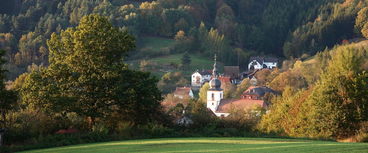 A rural landscape with a church, houses, and a forested hill.