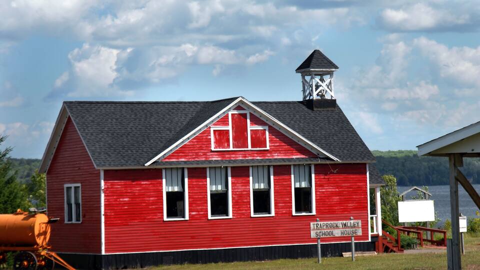 A red schoolhouse with a white trim roof and a bell tower, labeled 'Trappock Valley School House,' near a body of water.