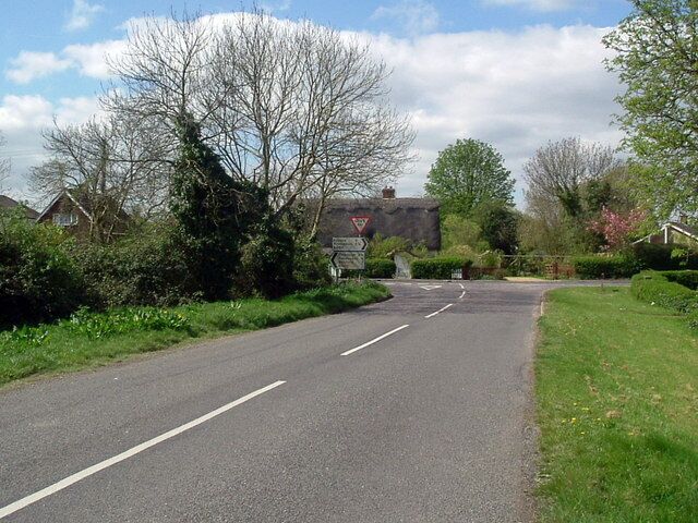 A rural road with a white dashed center line, surrounded by greenery and houses.