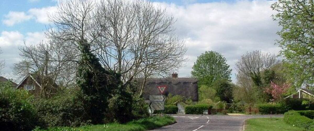 A rural road with a white dashed center line, surrounded by greenery and houses.