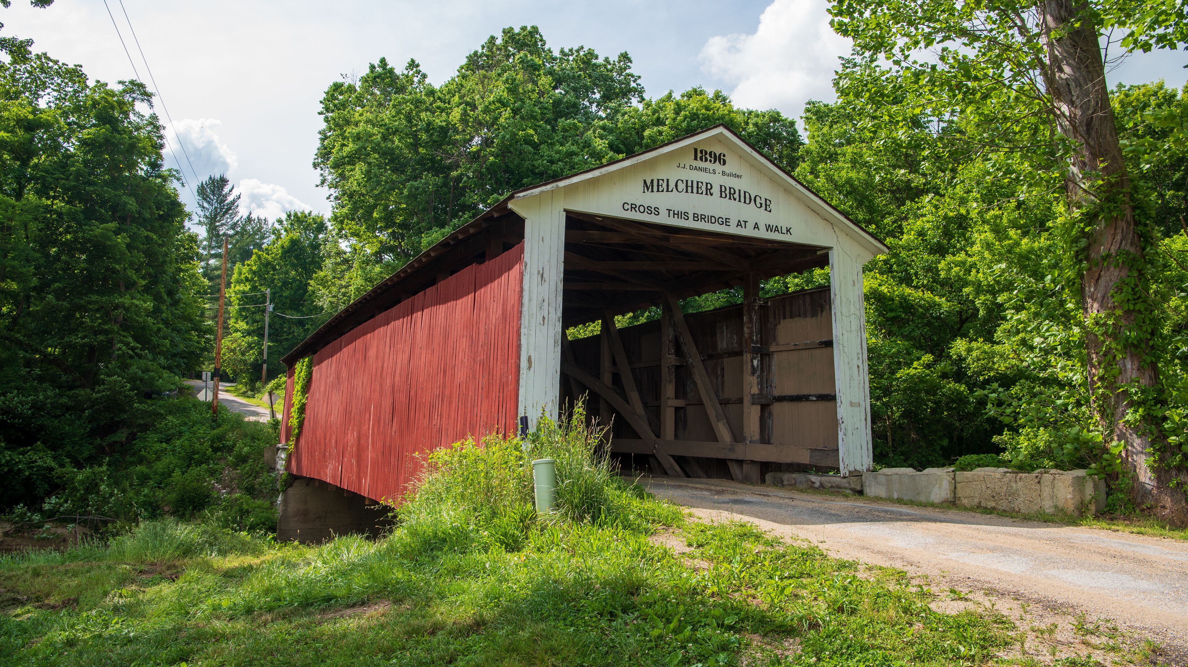 A red covered bridge with a white sign reading "1896 J.J. Daniels - Builder MELCHER BRIDGE CROSS THIS BRIDGE AT A WALK."