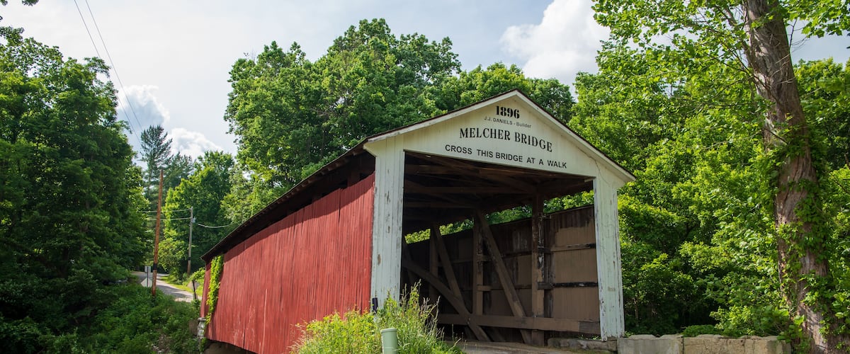 A red covered bridge with a white sign reading "1896 J.J. Daniels - Builder MELCHER BRIDGE CROSS THIS BRIDGE AT A WALK."