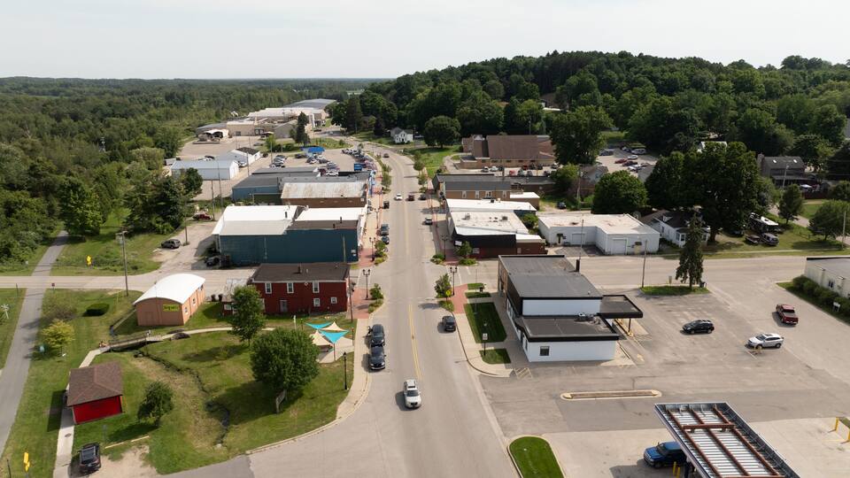 An aerial view of a small town with a main road, industrial buildings, and surrounding greenery.