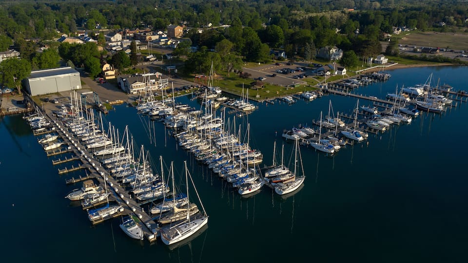 An aerial view of a marina with numerous boats docked in calm waters, surrounded by a small town and greenery.
