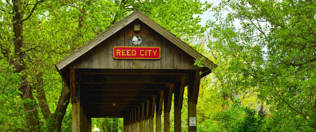 A wooden covered bridge with a sign reading "Reed City" in a lush green forest.