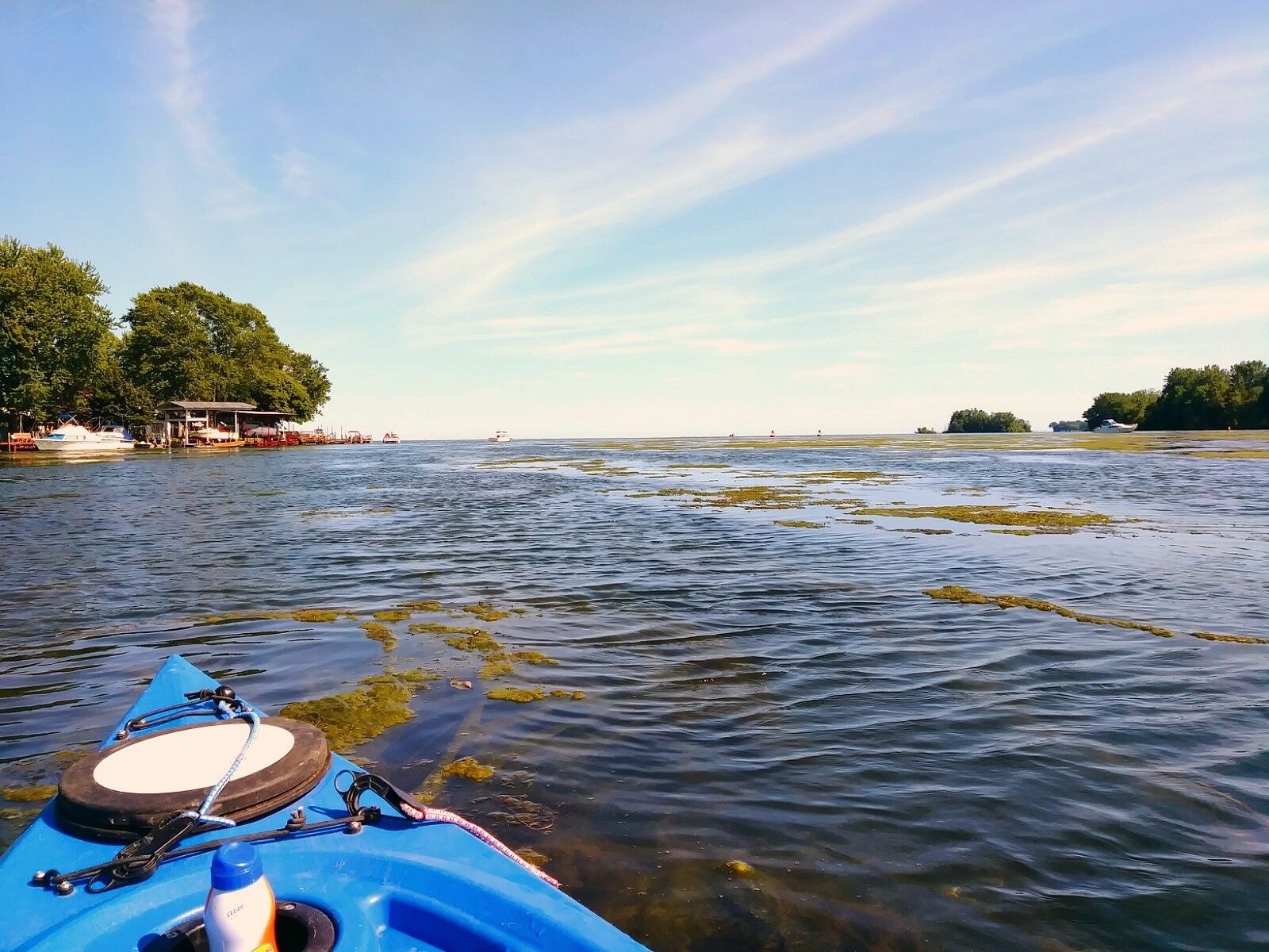 A calm lake with a blue kayak in the foreground, surrounded by trees and distant boats.