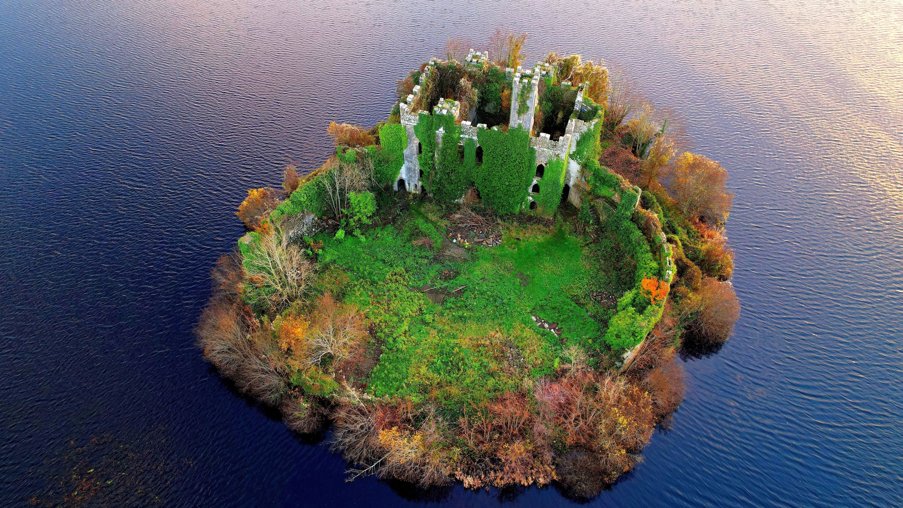An aerial view of a circular island with a central ruin surrounded by dense vegetation and a body of water.