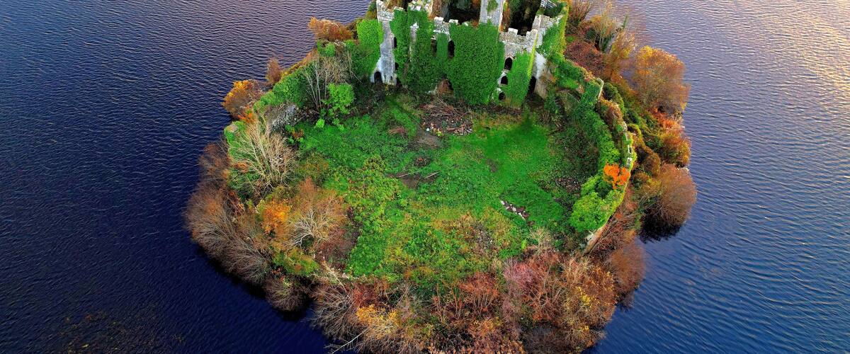 An aerial view of a circular island with a central ruin surrounded by dense vegetation and a body of water.