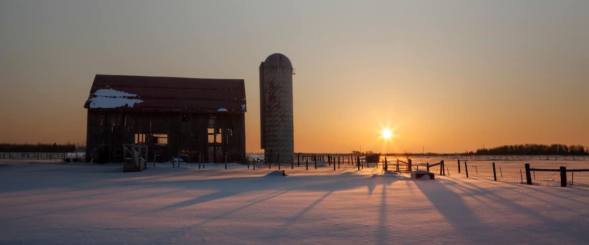 A snow-covered landscape with a barn and silo during sunset.