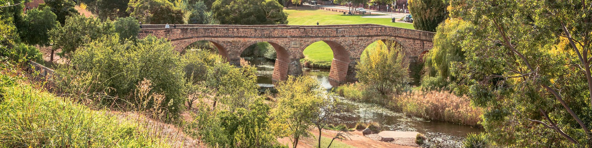 A stone bridge over a river, surrounded by trees and a park with a distant church spire.