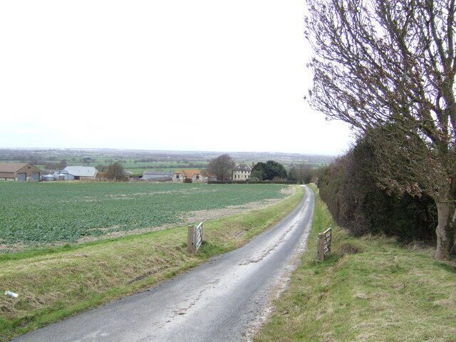 A rural road curving through fields with farm buildings in the distance.